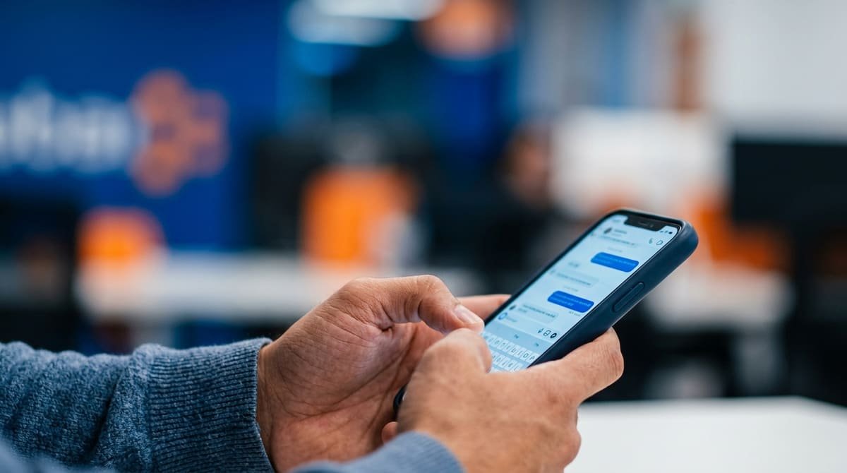 Close em mãos segurando um smartphone moderno e digitando agilmente na tela contra um fundo desfocado de escritório.